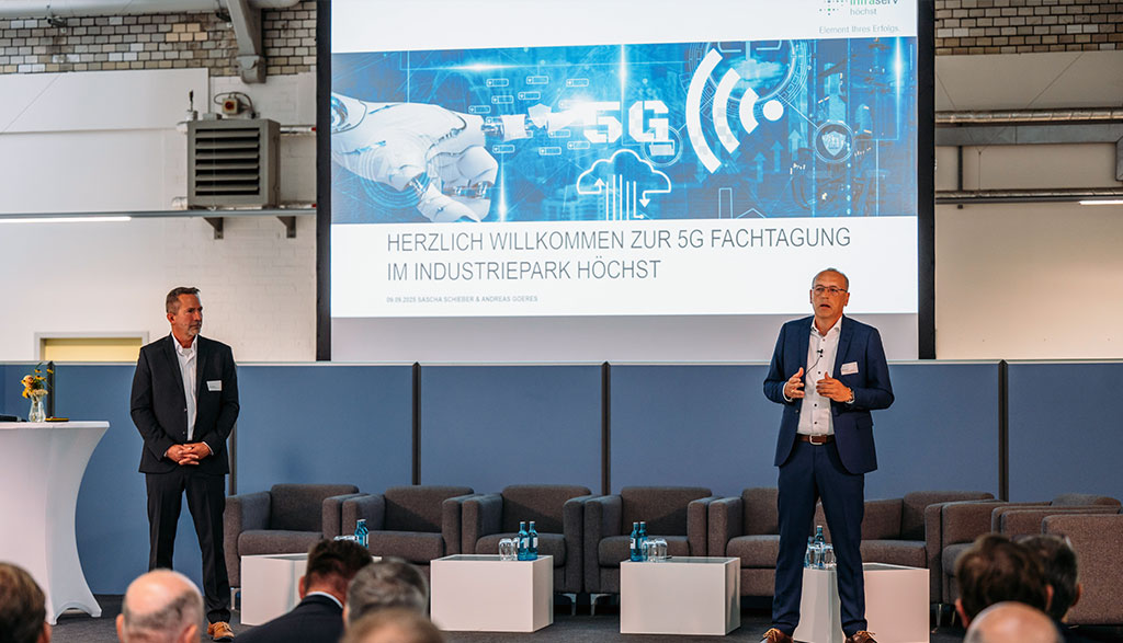 Zu sehen sind zwei Herren auf einer Bühne mit einem Präsentationsscreen im Hintergrund, mit dem Titel „Herzlich Willkommen zur 5G Fachtagung im Industriepark Höchst“