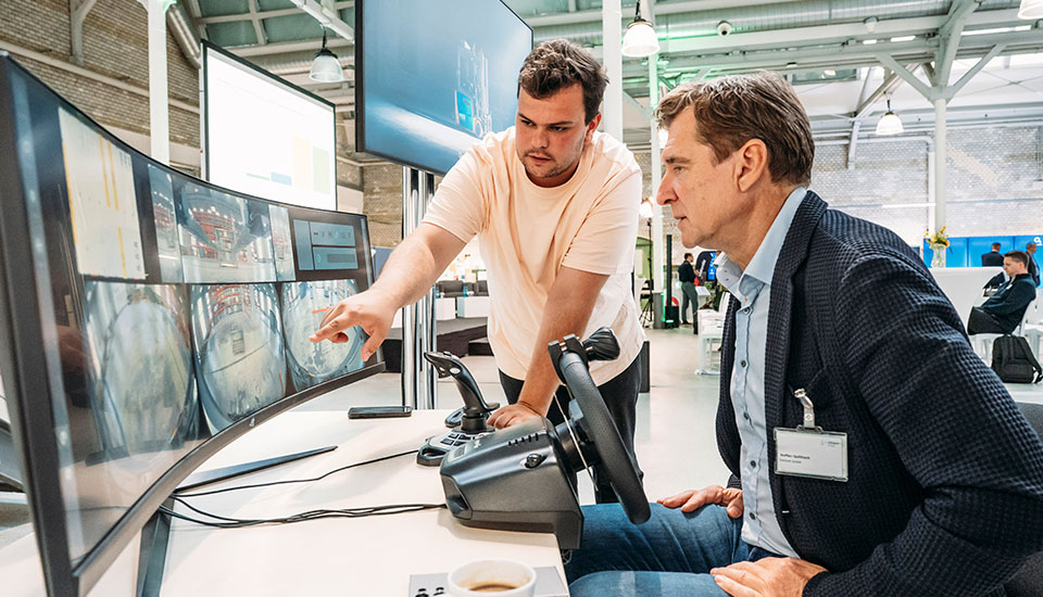 Zwei Personen demonstrieren auf der 5G-Fachtagung am Stand von enabl Technologies die Fernsteuerung eines Gabelstaplers über Monitore und Steuerpult.