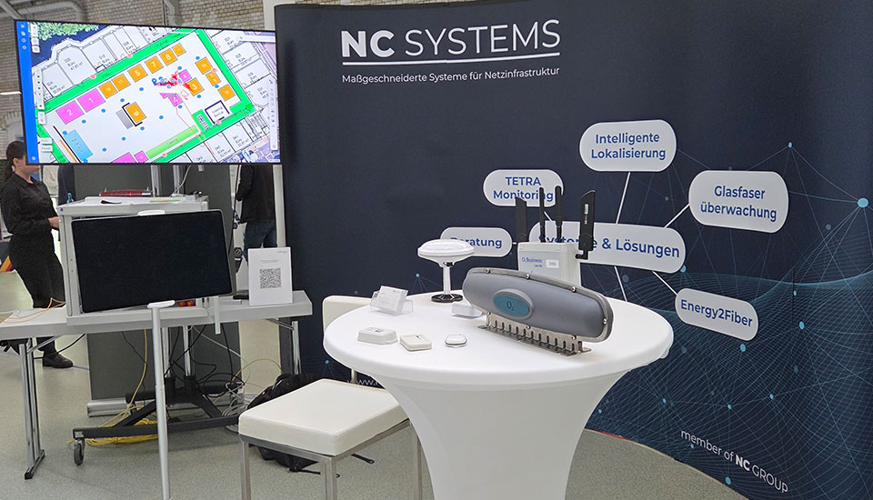 Messestand von NC Systems auf der 5G-Fachtagung mit Monitor, Infowand und Exponaten zu intelligenter Lokalisierung, Glasfaserüberwachung und Netzinfrastruktur.