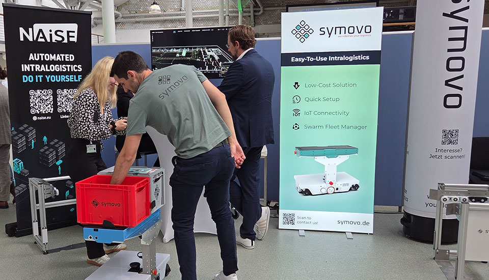 Messestand von Symovo und Naise auf der 5G-Fachtagung: Vorführung eines autonomen Intralogistik-Roboters mit Transportbox, ergänzt durch Infodisplays und Banner.