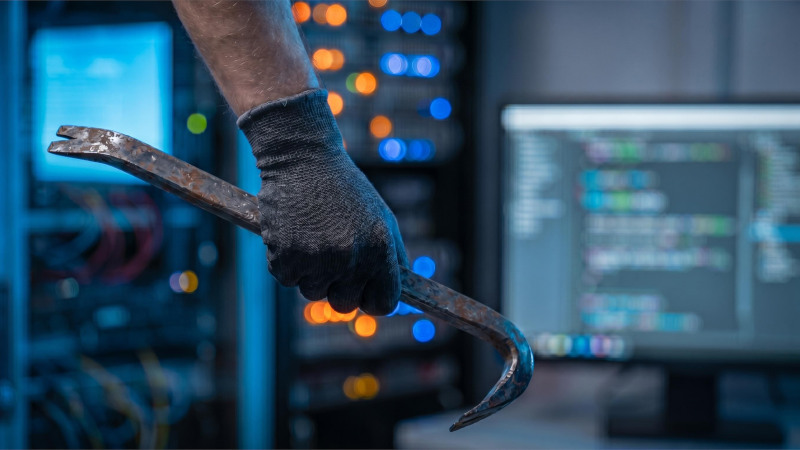 Symbolbild: Eine Hand hält ein Brecheisen, im Hintergrund sind Server mit leuchtenden LEDs und ein Computer zu sehen.