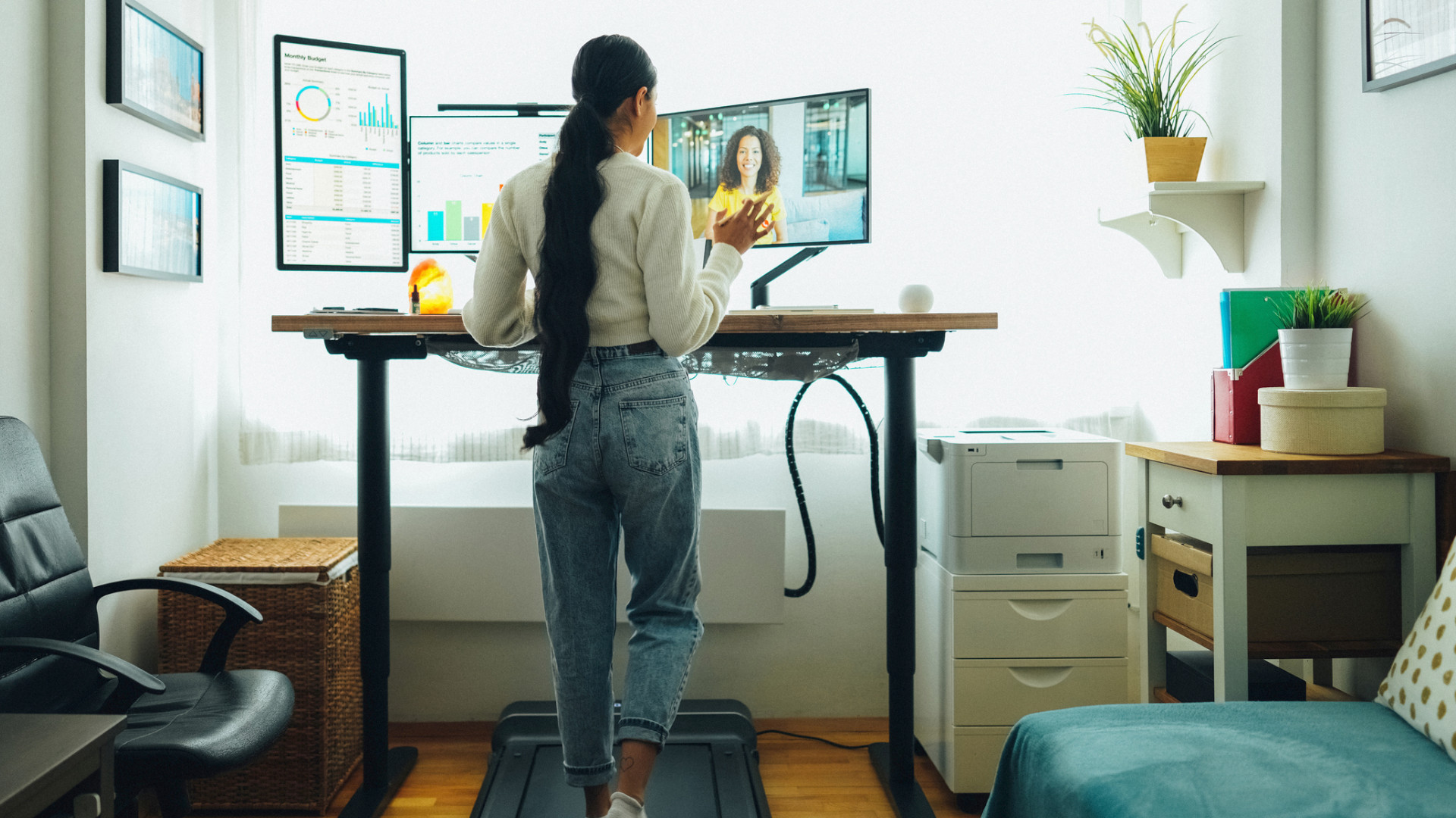 Junge Frau arbeitet im Homeoffice an einem Stehschreibtisch mit mehreren Monitoren und geht dabei auf einem Laufband.