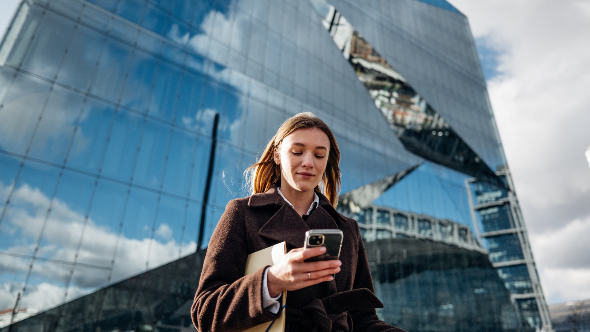 Eine junge Frau vor einem Hochhaus mit gläserner Fassade schaut auf ihr Handy