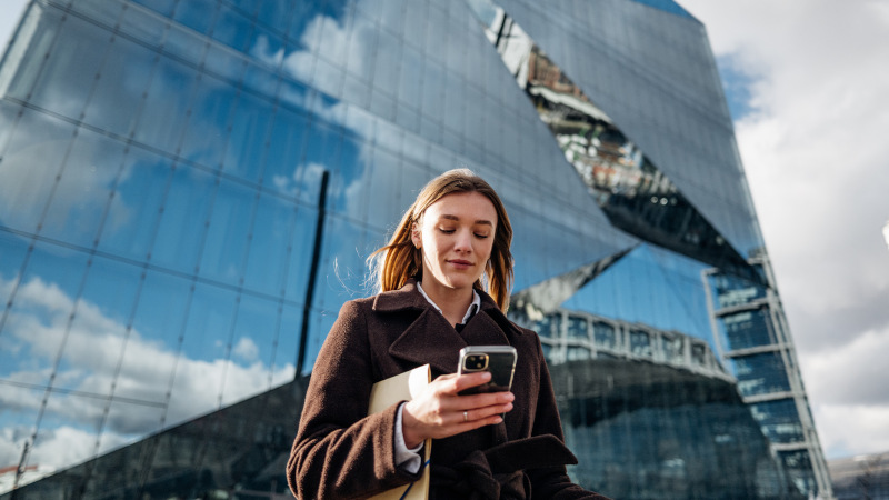 Eine junge Frau vor einem Hochhaus mit gläserner Fassade schaut auf ihr Handy