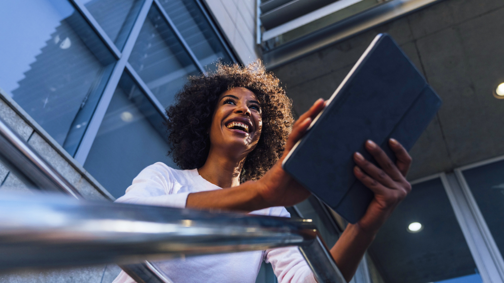 Abbildung: Eine Person steht im Freien vor einem modernen Gebäude und hält ein Tablet mit beiden Händen. Die Person hat dunkles, lockiges Haar in mittlerer Länge und trägt ein langärmliges, weißes Oberteil. Die Perspektive ist von unten aufgenommen, sodass ein Metallgeländer im Vordergrund sichtbar ist. Im Hintergrund sind Glas- und Betonfassaden sowie Lamellenstrukturen zu erkennen. Die Szene ist bei Tageslicht aufgenommen und vermittelt einen urbanen, technologischen Kontext.