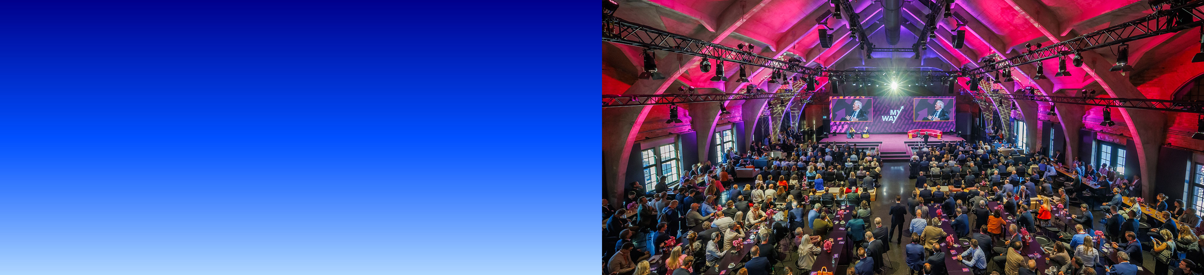Historische Gewölbehalle mit Traversen und pinker Beleuchtung, gefüllt mit Gästen an langen Tischen vor einer Bühne