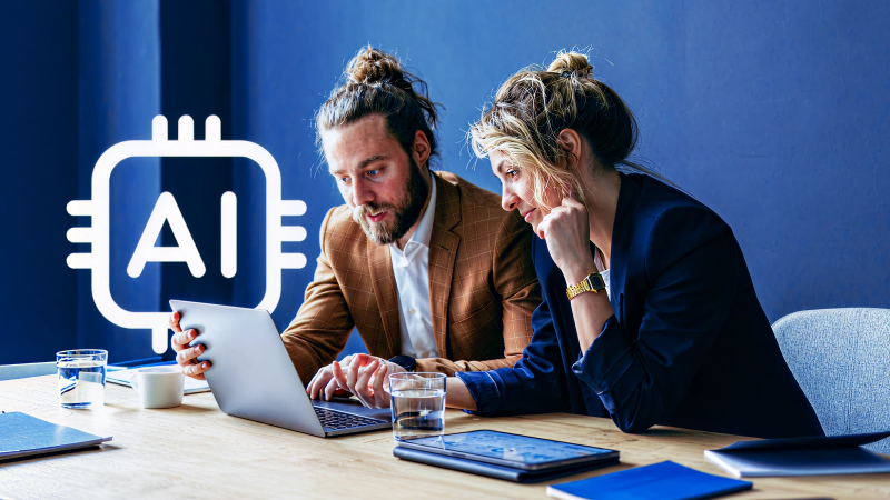 Abbildung: Eine Frau und ein Mann sitzen vor einem Laptop, im Hintergrund ist ein weißes Icon mit dem Schriftzug AI zu sehen