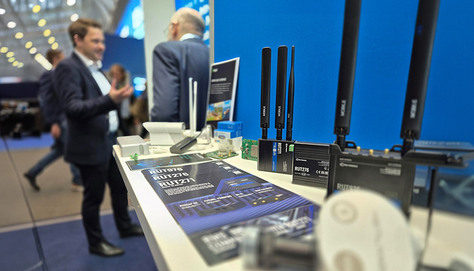 Messestand mit ausgestellten Mobilfunk-Routern und Antennen (z. B. RUT276) auf einem Tisch; im Hintergrund Besucher im Gespräch.