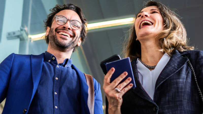 Abbildung: Zwei junge Erwachsene stehen in einem modernen Innenraum.  Linke Person (männlich): trägt ein blaues Sakko über einem dunklen Hemd, hat kurze, leicht gewellte Haare, trägt eine Brille und eine braune Umhängetasche über der Schulter. Rechte Person (weiblich): trägt ein dunkles Business-Jacket über einer weißen Bluse, hat schulterlanges, gewelltes Haar und hält ein Smartphone sowie ein kleines Notizbuch in der Hand. Beide lachen.  Im Hintergrund sind metallische Strukturen und Lichtleisten zu sehen. Die Szene vermittelt einen professionellen, geschäftlichen Kontext.