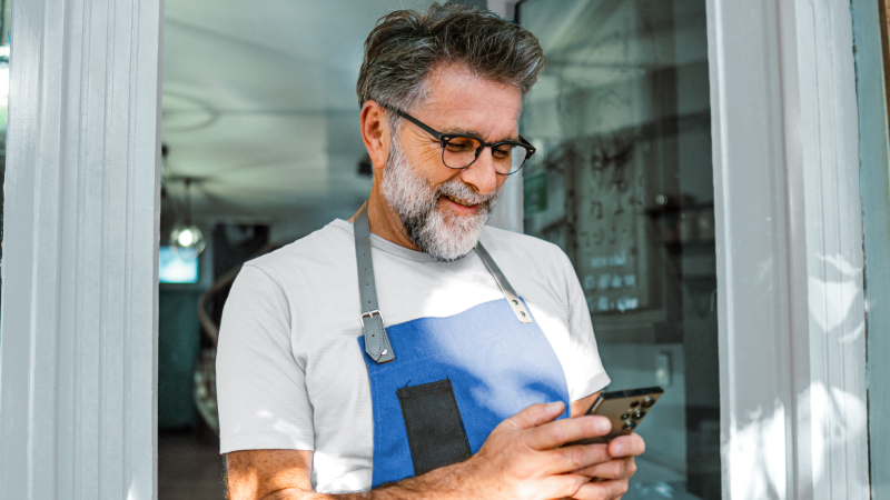 Abbildung: Älterer Mann mit Brille und Bart in blauem Arbeitskittel schaut auf sein Smartphone und steht in einer Türöffnung.