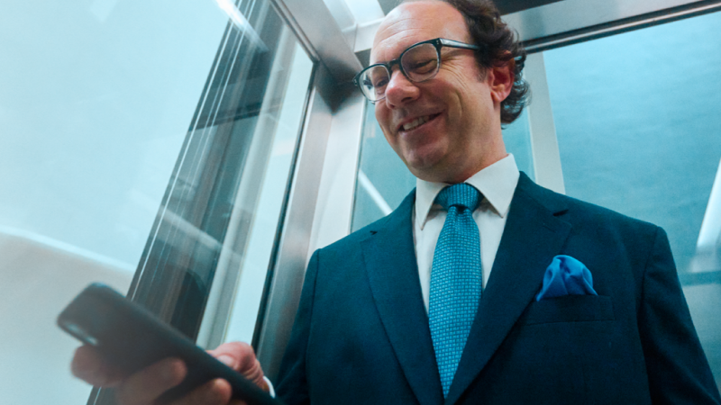 Abbildung: Mann mittleren Alters mit Halbglatze und Brille im dunkelblauen Anzug mit Krawatte und Einstecktuch sieht lächelnd auf das Smartphone in seiner Hand
