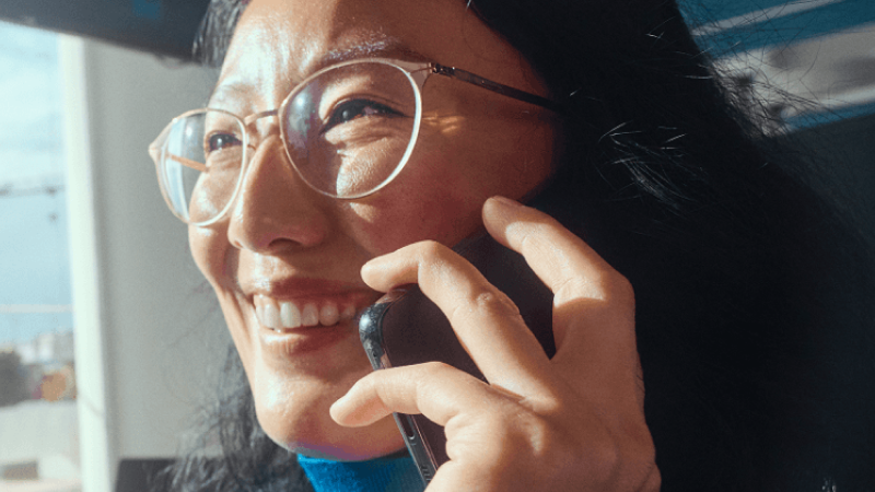 Abbildung: Frau mit dunklen Haaren brauner Haut und Brille hält strahlend Handy ans Ohr