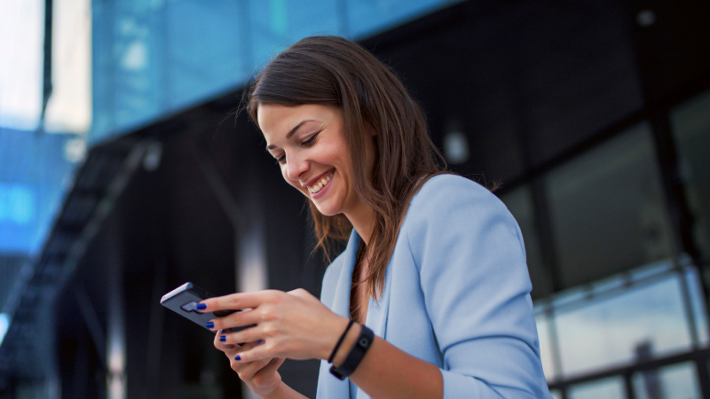 Abbildung: Eine junge Frau steht vor einem modernen Glasgebäude und hält ein Smartphone in beiden Händen, als würde sie etwas darauf prüfen.  Die Person trägt ein hellblaues Sakko über einem dunklen Oberteil und ein Fitnessarmband am linken Handgelenk. Die Haare sind lang und glatt, fallen über die Schultern.  Im Hintergrund sind Glasfassaden und metallische Strukturen zu sehen. Die Szene vermittelt einen urbanen, professionellen Kontext.