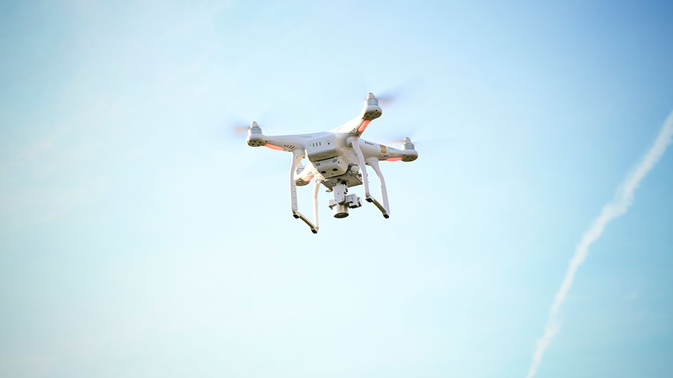 Weiße Drohne mit Kamera fliegt vor blauem Himmel mit Kondensstreifen